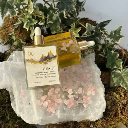 Two gold and white Heart Body Oil  bottles on a rock with pink crystals and greenery