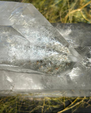 A close-up of a clear quartz crystal point with natural inclusions, resting outdoors on a bed of dry grass.