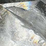A close-up of a translucent clear crystal or gemstone with sharp facets, natural inclusions, and a subtle iridescent rainbow sheen under light.