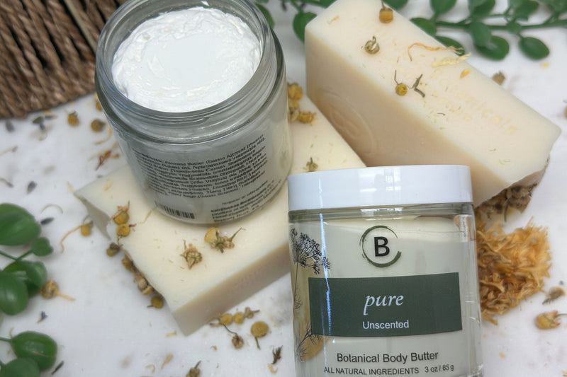 Two jars of Botanical Body Butter with bars of soap on a textured surface with greenery.