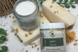 Two jars of Botanical Body Butter with bars of soap on a textured surface with greenery.