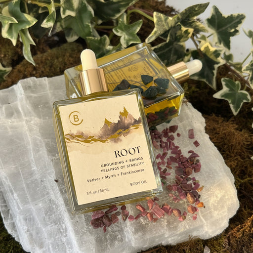 Two bottles of 'Root' body oil on a textured stone surface with plants in the background