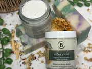 Two jars of 'ultra calm' botanical body butter with dried herbs and soap on a marble surface.