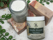 Two jars of 'vanilla blossoms' botanical body butter and a bar of soap on a textured surface with greenery.